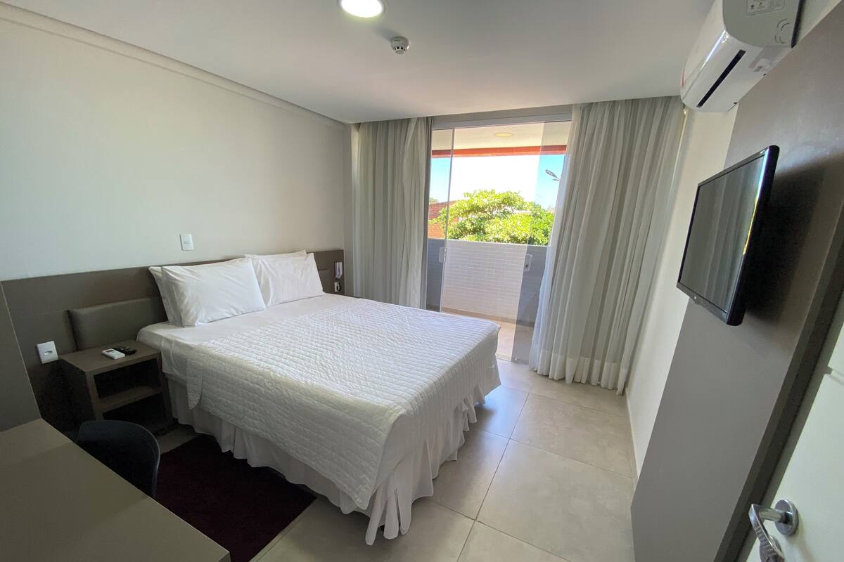 A hotel room with a bed, a desk, a chair, a TV, and a balcony with curtains.