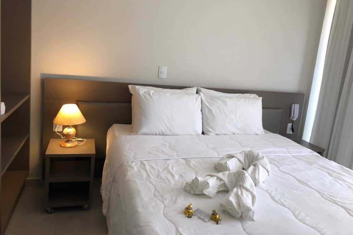 A hotel room with a neatly made bed, a bedside lamp, a phone, and a shelf.
