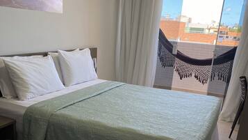 Habitación de hotel con cama, almohadas blancas, colcha verde, una mesita pequeña con aperitivos, alfombra oscura y vista a edificios a través de una ventana con cortinas.