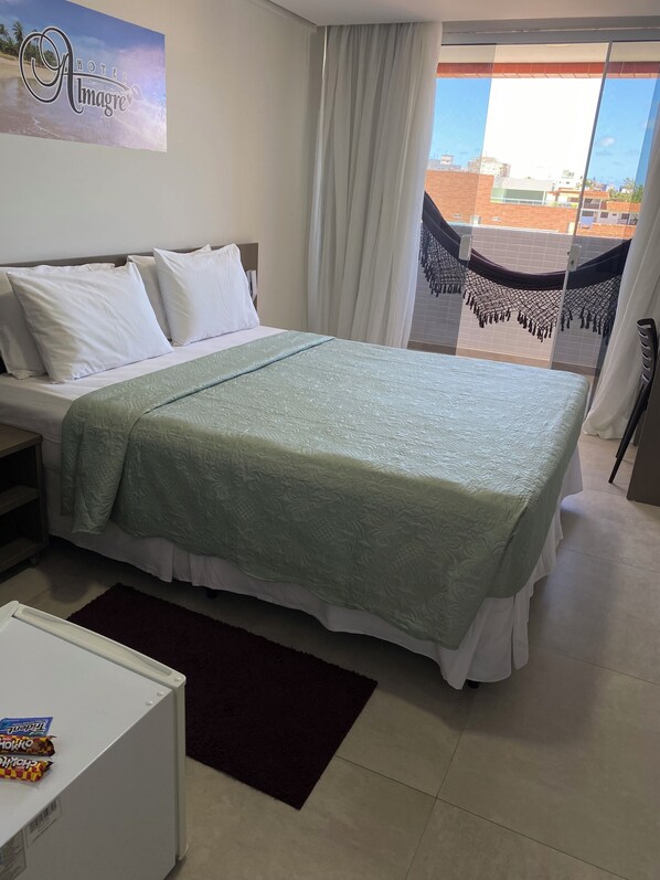 Habitación de hotel con cama, almohadas blancas, colcha verde, una mesita pequeña con aperitivos, alfombra oscura y vista a edificios a través de una ventana con cortinas.