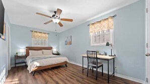 Ein Schlafzimmer mit Bett, Schreibtisch, Stuhl, Deckenventilator und einem Fenster mit Vorhängen.