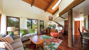 Amplia sala de estar con techo de madera, un sofá con cojines verdes, una mesa de centro de madera y vistas a un jardín exuberante a través de grandes ventanales.