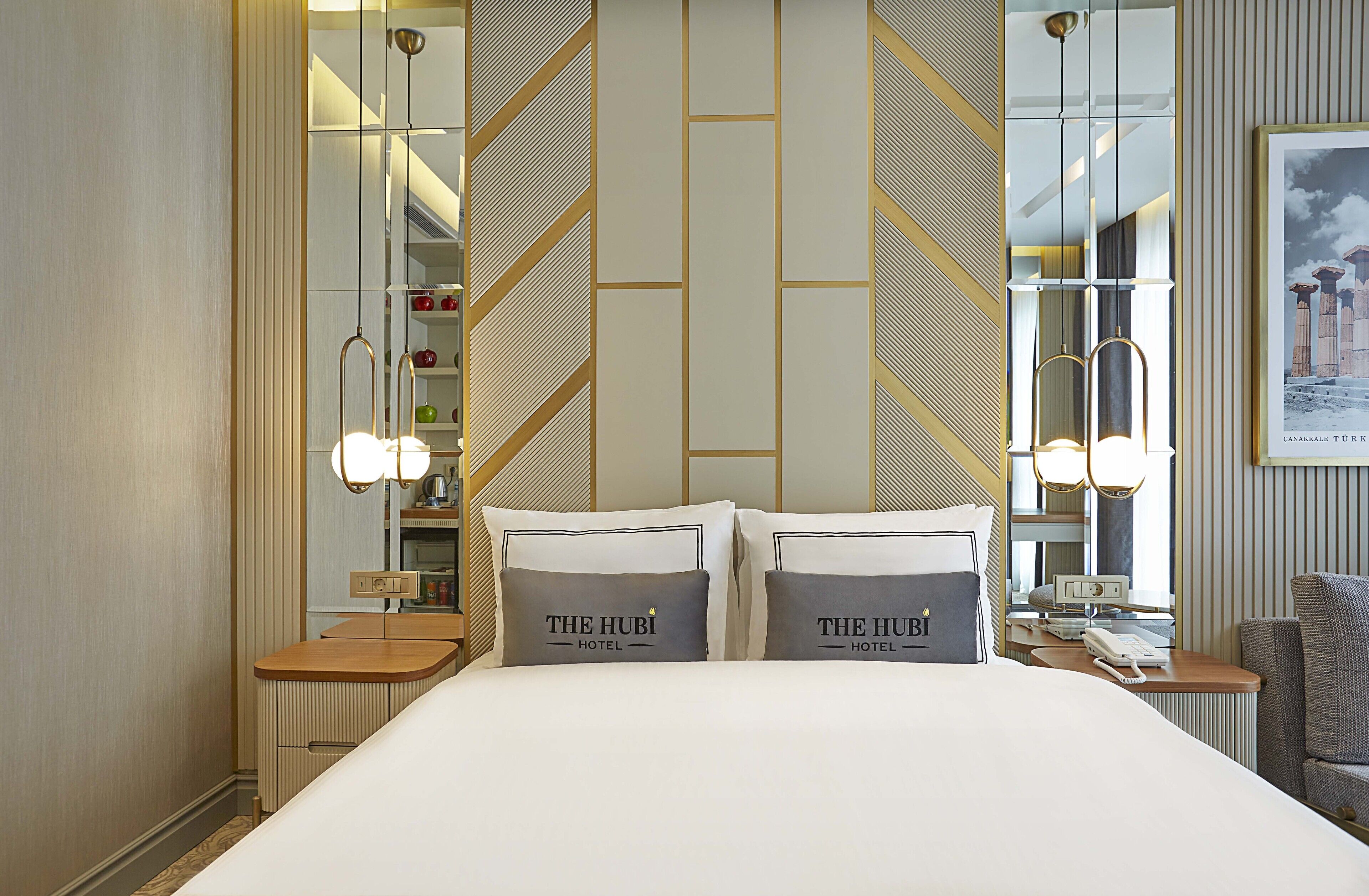 A hotel room with a large bed, two pillows with the hotel's name, a bedside table with a lamp, a mirror, and a framed picture on the wall.