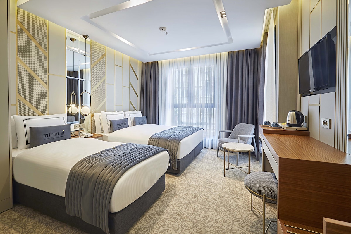 A hotel room with two beds, a desk, a chair, a TV, and a large window with curtains.