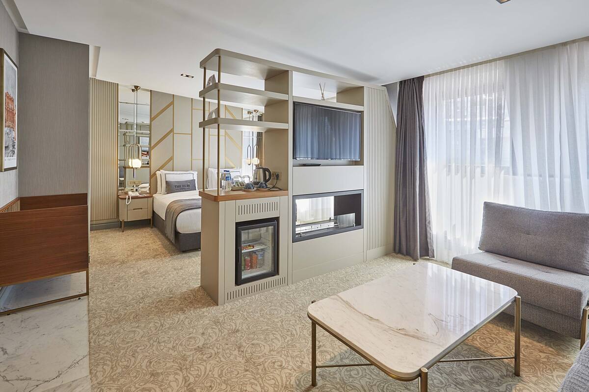 A modern hotel room with a bed, a sofa, a TV, a minibar, and a large window with curtains.