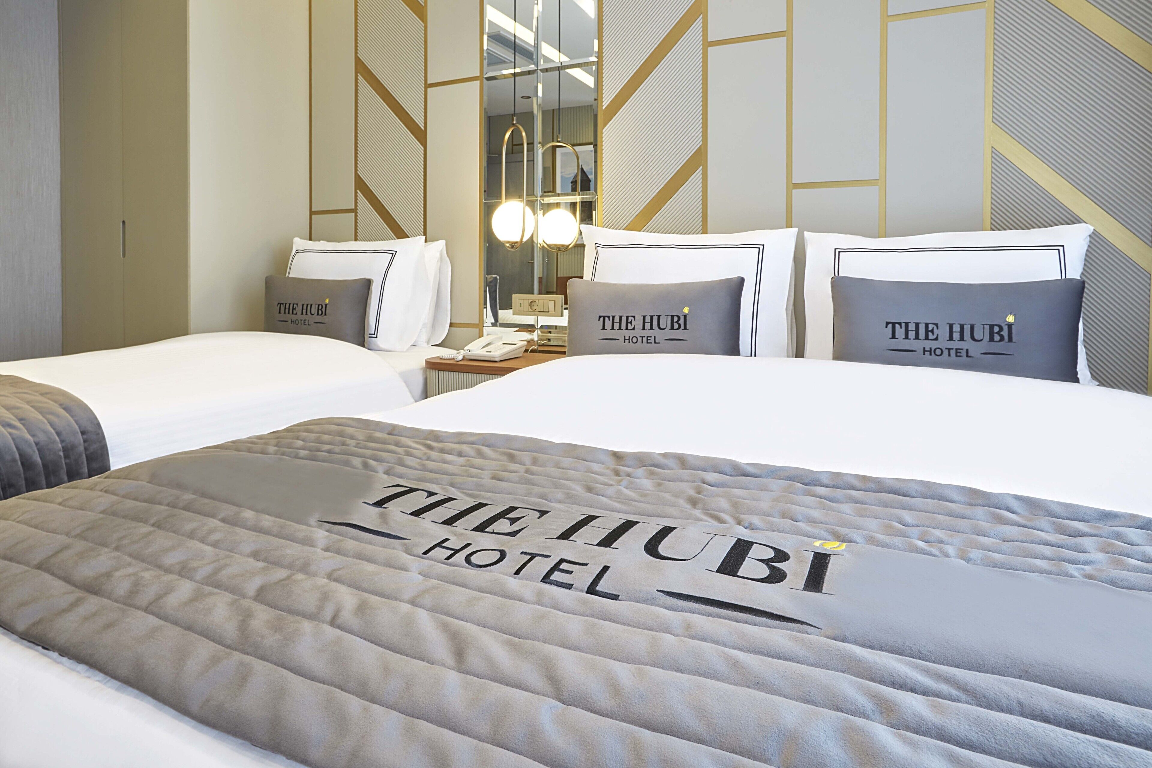 Habitación de hotel con una cama grande, dos almohadas con el logotipo del hotel, una mesita de noche con una lámpara y un espejo.
