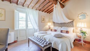 A bedroom with a large bed, a bench, a bedside table with lamps, and a mirror on the wall.