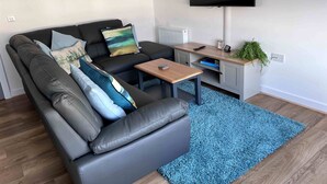 A modern living room with a grey leather sofa, a blue rug, a wooden coffee table, a TV mounted on the wall, and a small plant on a wooden shelf. - Caledonia Place - Retreat Group (Aviemore)