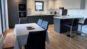 A modern kitchen with a dining area, featuring a central island, built-in appliances, and a window with blinds. - Caledonia Place - Retreat Group (Aviemore)