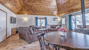 Ein gemütliches Blockhäuschen-Interieur mit Holzbalkendecke, einem Wohnbereich mit Ledersofas, einem runden Esstisch und einem großen Fenster mit Blick auf schneebedeckte Bäume.