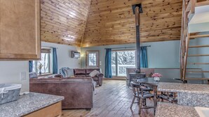 Ein gemütliches Blockhaus-Interieur mit Holzbalkendecke, Kamin, Essbereich und Wohnzimmer mit Sofa und Sesseln.