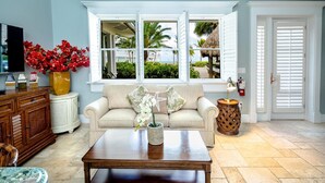 A living room with a beige sofa, a wooden coffee table, a TV on a wooden cabinet, and a window with a view of a beach and palm trees. - 1 bedroom, 1 bath, full kitchen, 1 queen bed, 1 queen sleeper sofa. (Islamorada)