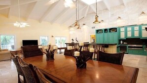A spacious dining area with a large wooden table and multiple chairs, a kitchen with teal cabinets, and a high ceiling with exposed beams. - Jamaican Palm House 3 Bedrooms, 2 Bath, Poolside (Islamorada)