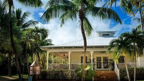 A white building with a covered porch, surrounded by palm trees and a clear sky. - Jamaican Palm House 3 Bedrooms, 2 Bath, Poolside (Islamorada)
