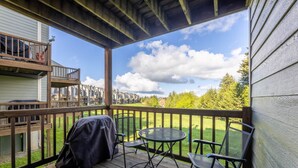 Eine Holzterrasse mit Grill und zwei Stühlen, mit Blick auf eine grüne Landschaft und eine Häuserreihe.