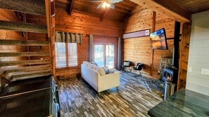 A cozy cabin interior with a wooden staircase, a sofa, a stone fireplace, and a television mounted on the wall.