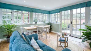 Une pièce lumineuse et ensoleillée, dotée d’une grande fenêtre, d’une table à manger en bois, de chaises blanches, d’un canapé bleu et d’une petite table ronde sur laquelle sont posés des livres et une tasse.