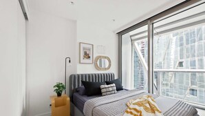 Een moderne slaapkamer met een groot raam, een bed met grijs hoofdeinde, een nachtkastje met een plant en een lijst met een afbeelding aan de muur.