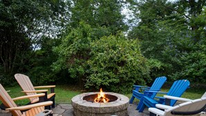 A circular fire pit surrounded by Adirondack chairs in a garden setting.
