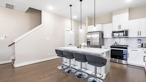 Une cuisine moderne avec des armoires blanches, des appareils en acier inoxydable, un îlot central doté de tabourets de bar et un escalier menant à l’étage supérieur.