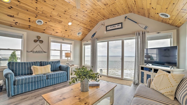 Una sala de estar acogedora con techo de madera, ventanas amplias con vistas a un cuerpo de agua, un televisor y una zona de estar confortable.