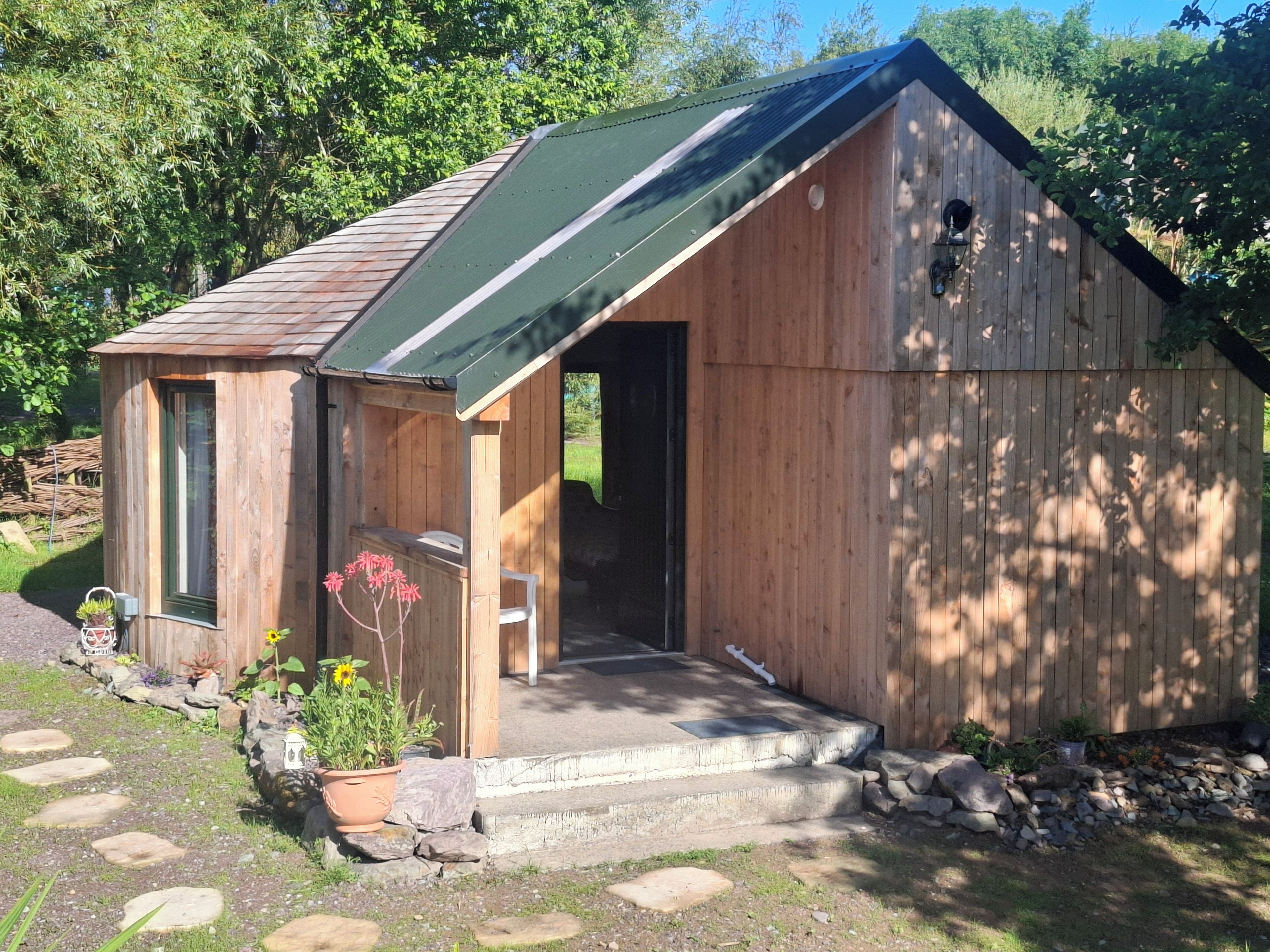 Une cabane en bois avec un toit vert, une véranda avec des marches et un petit jardin agrémenté de plantes et de pierres plates servant de pas.