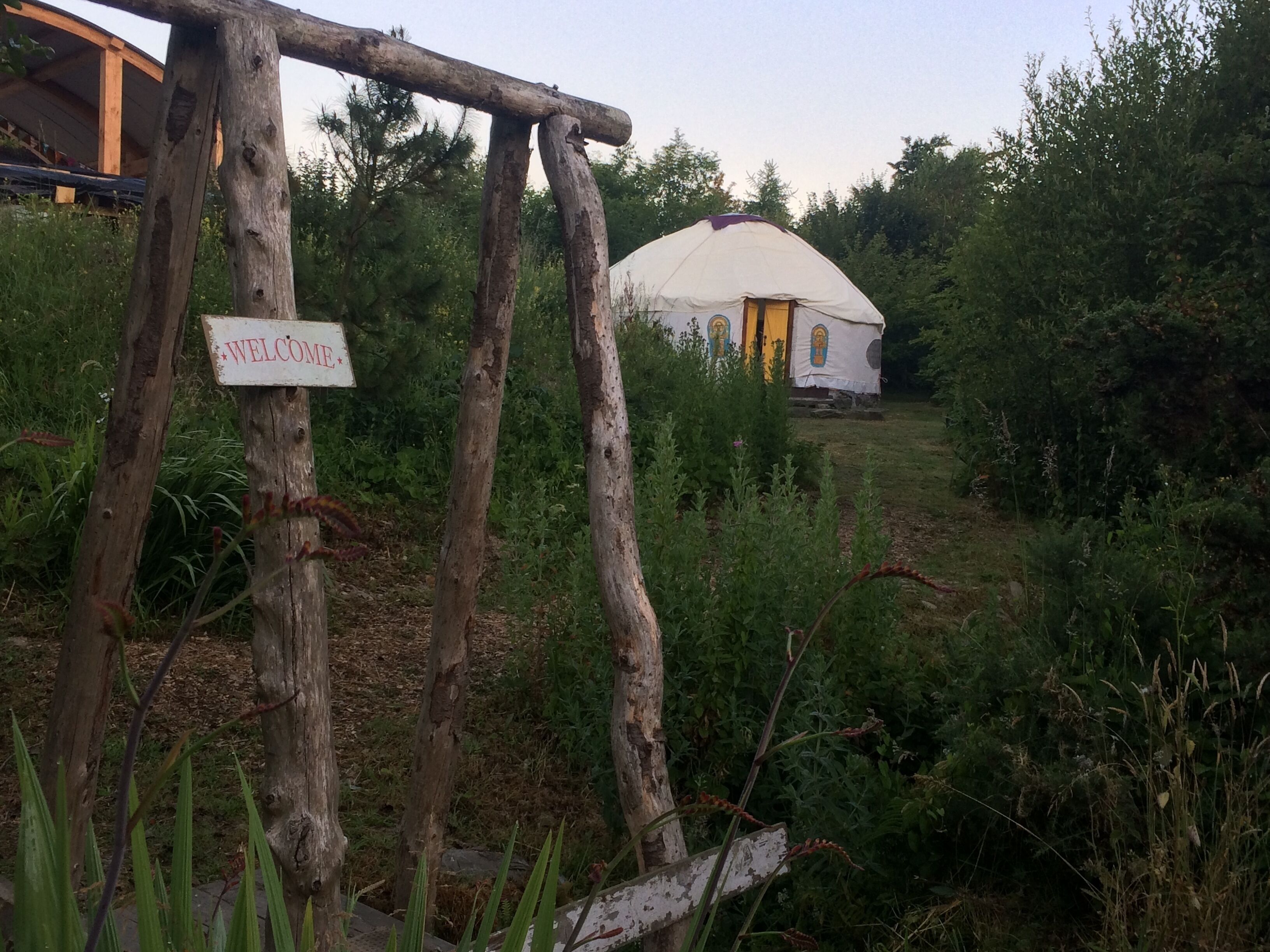 A rustic wooden archway with a 'WELCOME' sign, leading to a traditional yurt with a yellow door, surrounded by lush greenery.