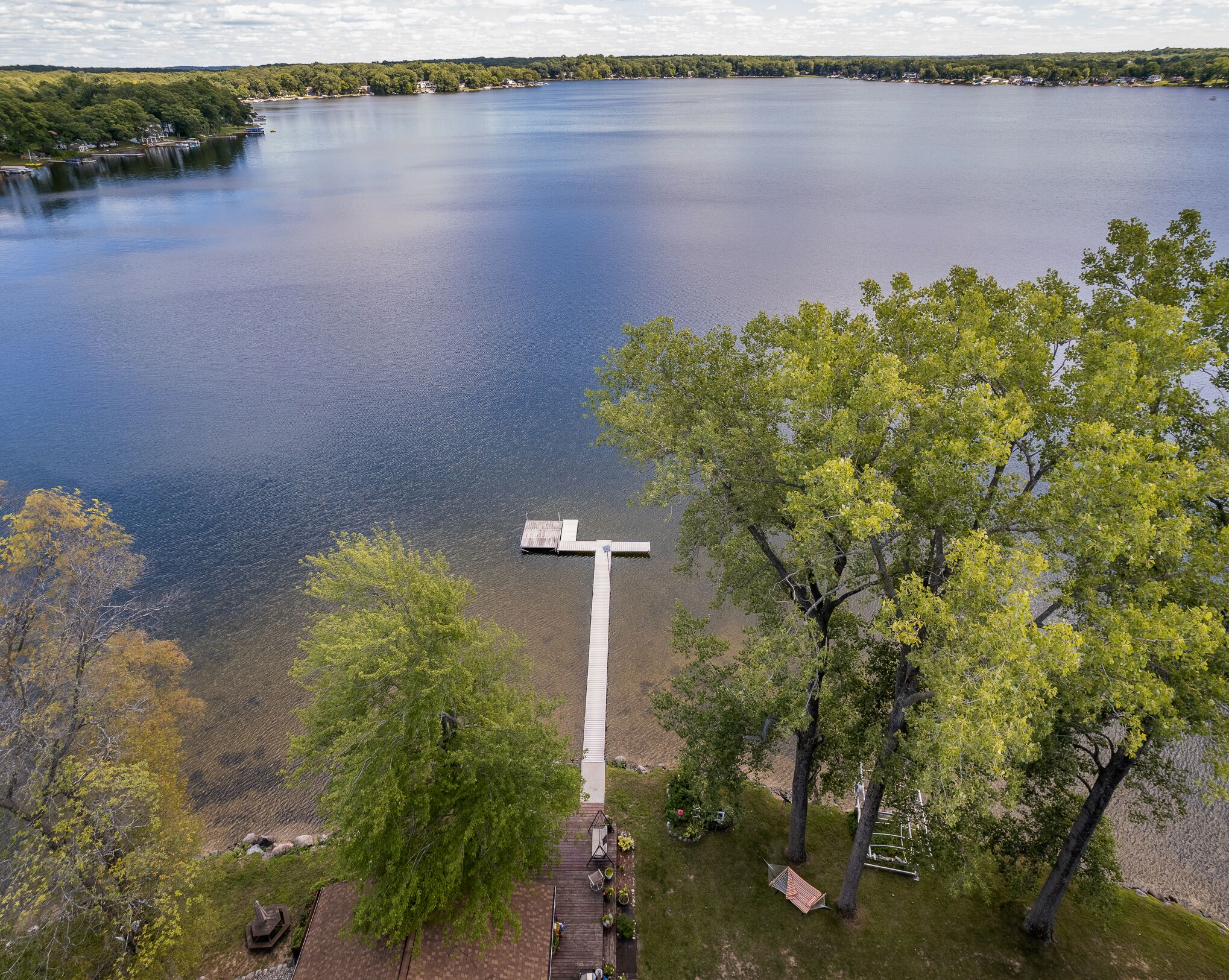 Vista de un lago con un muelle que se adentra en el agua, rodeado de árboles y un área verde con mobiliario exterior.