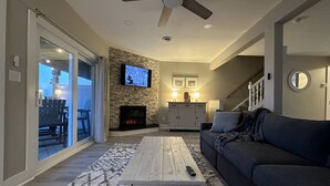 Una sala de estar con chimenea, un televisor montado en la pared, un ventilador de techo y un ventanal con vistas al exterior.