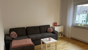 A modern living room with a dark grey sofa, a matching ottoman, and a small white coffee table. A pink throw blanket is draped over the ottoman. A white side table with a red book and a bowl is next to the sofa. The room has wooden flooring and a window with white curtains. - Oberursel old town very centrally located (Oberursel)