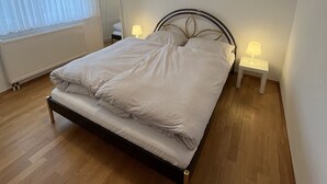 A neatly made bed in a room with a wooden floor, a small bedside table with a lamp, and a window with sheer curtains. - Oberursel old town very centrally located (Oberursel)