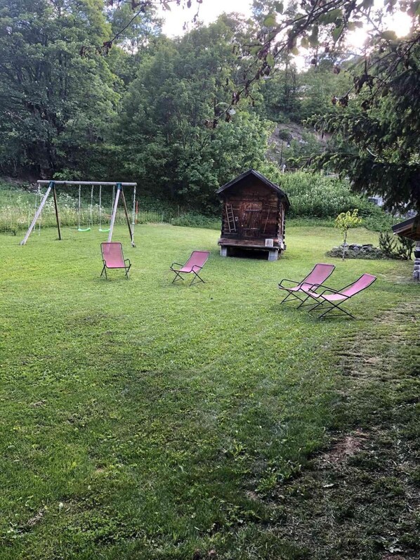 A grassy outdoor area with a wooden cabin, a swing set, and several lounge chairs.