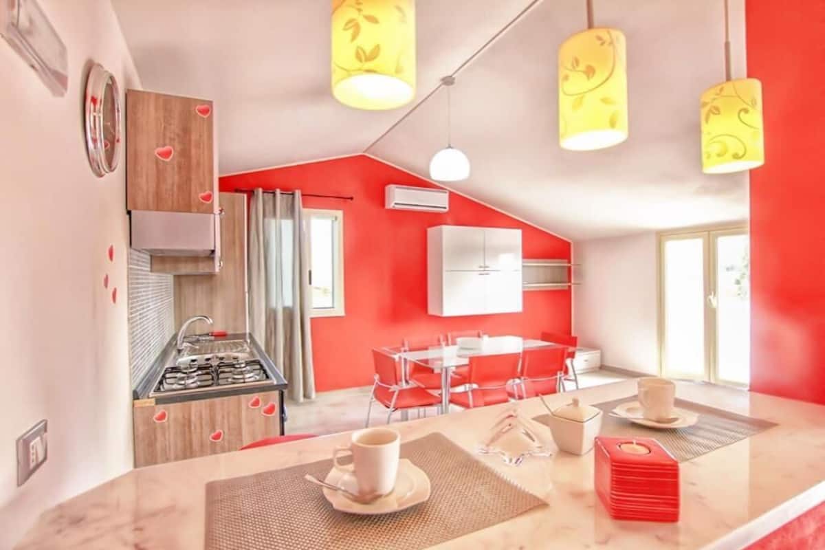 A modern kitchen with a red accent wall, a dining area with red chairs, and a bar area with a countertop and bar stools.