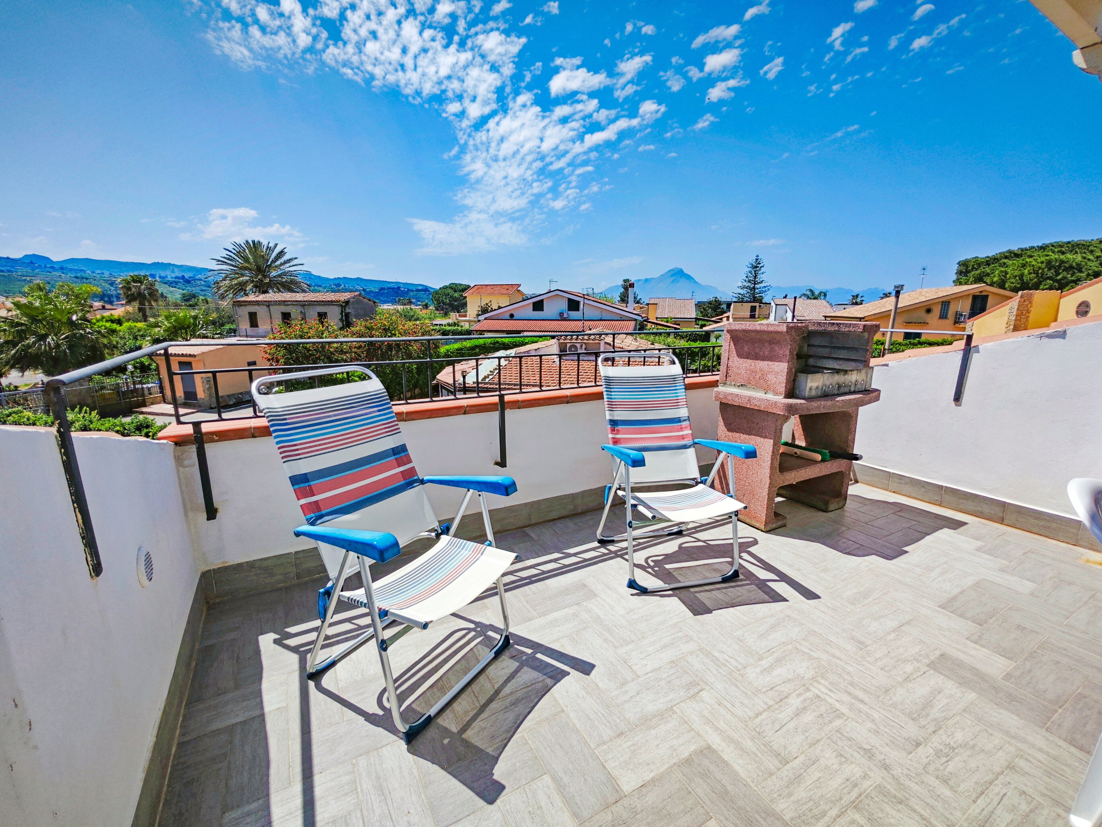 A terrace with two striped lounge chairs and a barbecue grill, overlooking a residential area with houses and a mountain in the background.