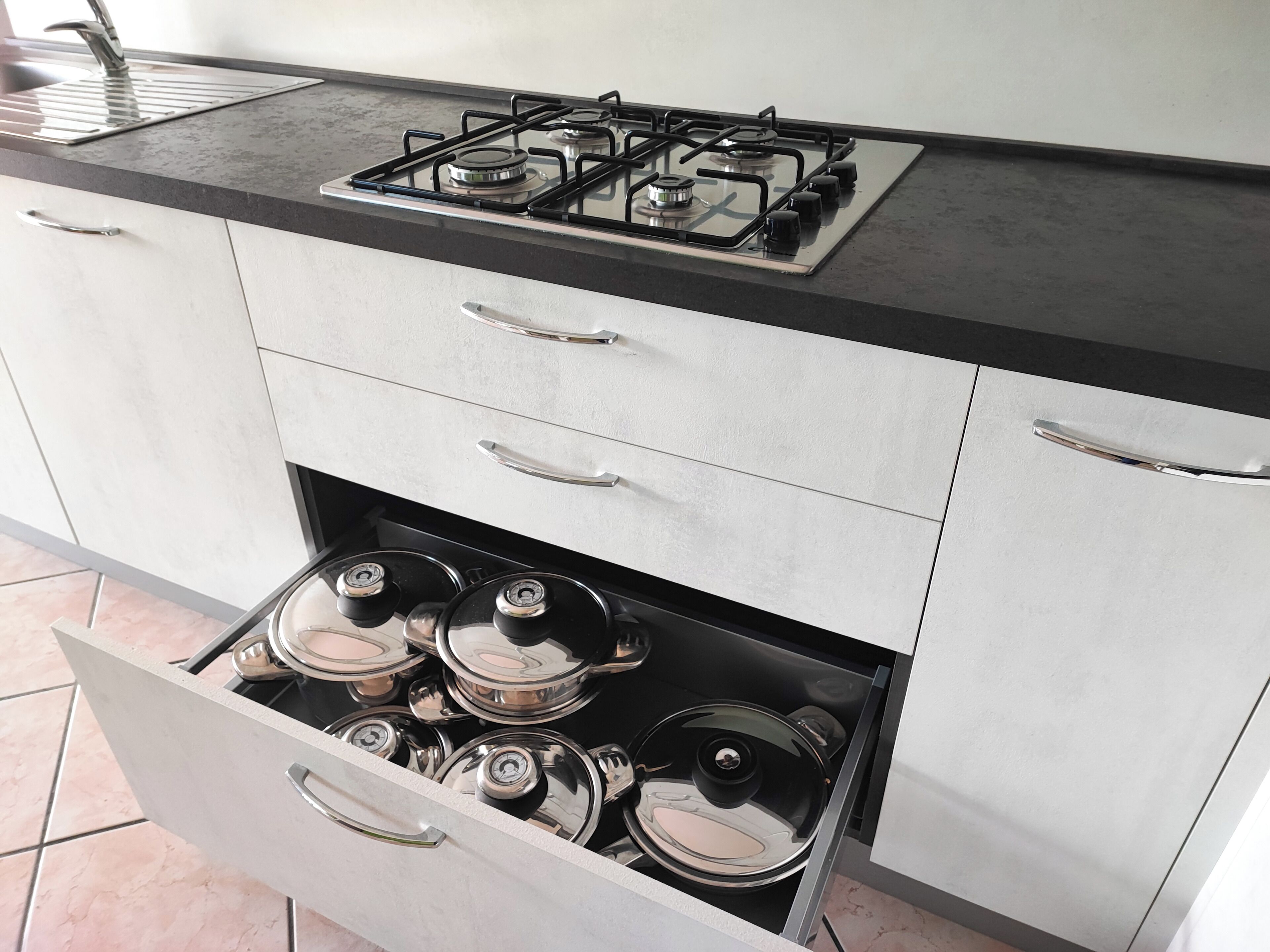 Una cocina con estufa a gas, ollas de acero inoxidable y un cajón con ollas.
