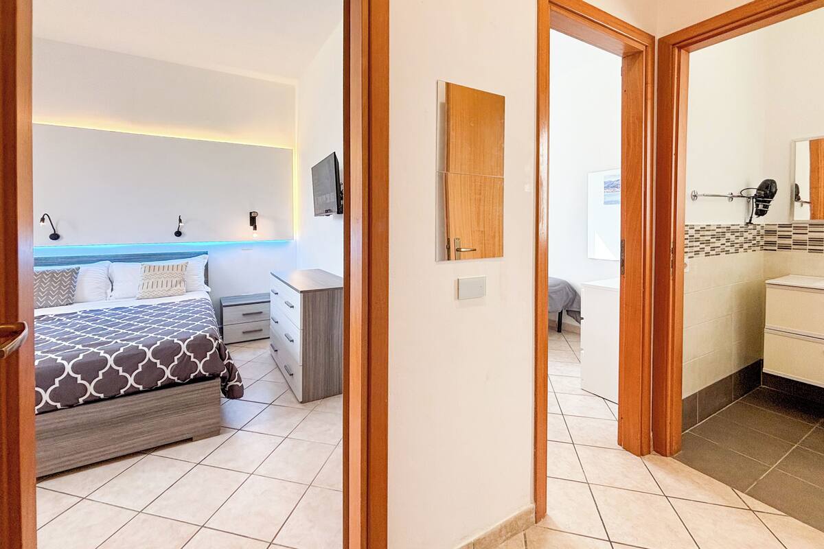 A bedroom with a bed, bedside table, and a nightstand. There is a bathroom with a sink and mirror.