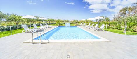 A rectangular swimming pool with white lounge chairs and umbrellas on either side, surrounded by a paved deck and greenery.