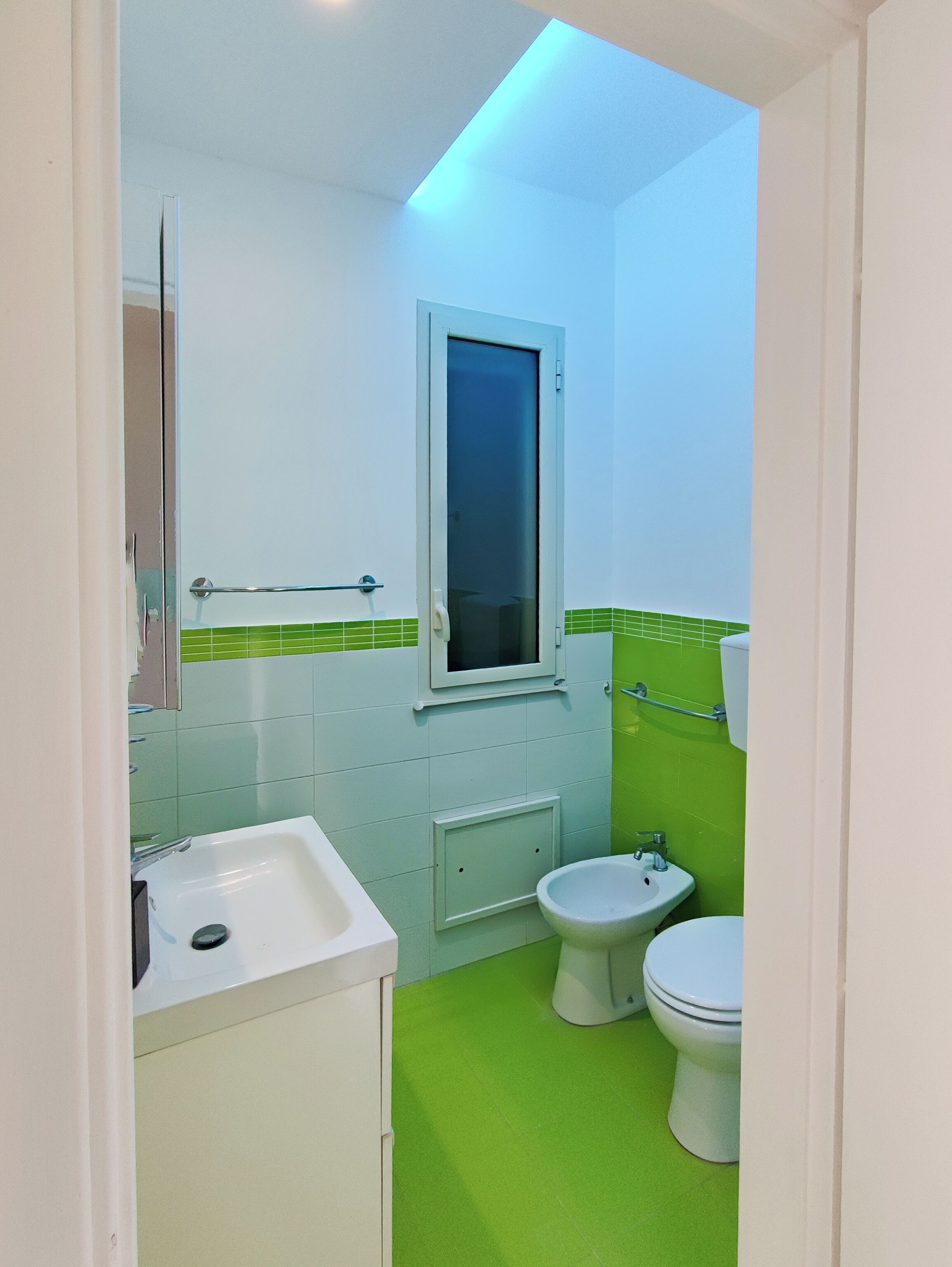 Une salle de bain avec un lavabo blanc, un bidet et des toilettes. Il y a une fenêtre au-dessus du lavabo et un mur recouvert de carreaux verts.