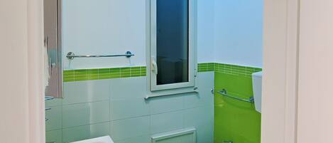 Une salle de bain avec un lavabo blanc, un bidet et des toilettes. Il y a une fenêtre au-dessus du lavabo et un mur recouvert de carreaux verts.