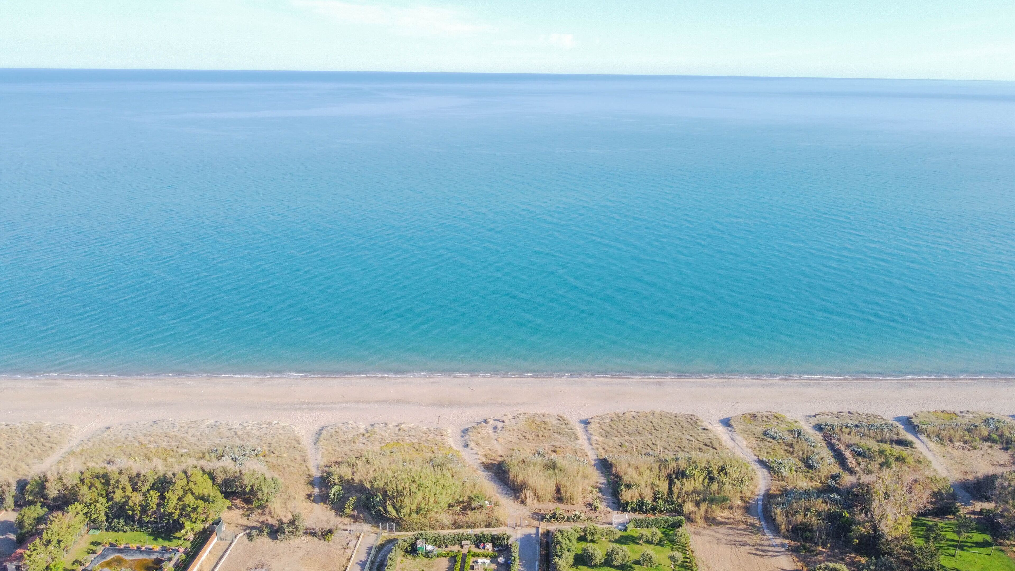 An aerial view of a coastal area with a beach and a road leading to a residential area with houses and greenery.