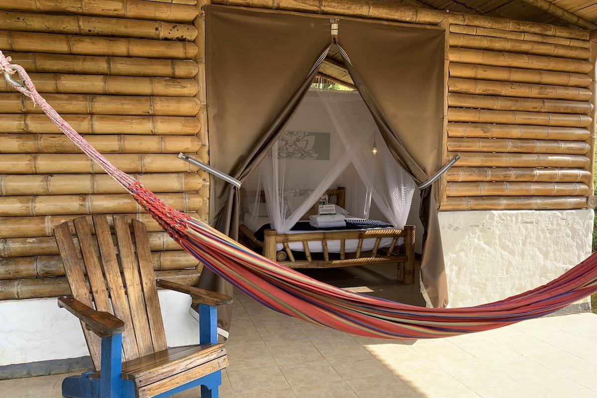 Una hamaca atada entre dos postes de madera, un banco de madera y una cama con mosquitera en una cabaña de bambú.
