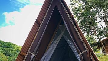 Una tienda de campaña triangular con una cama en su interior, montada sobre una plataforma de madera en una zona cubierta de césped, con árboles al fondo.
