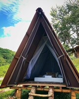 Una tienda de campaña triangular con una cama en su interior, montada sobre una plataforma de madera en una zona cubierta de césped, con árboles al fondo.