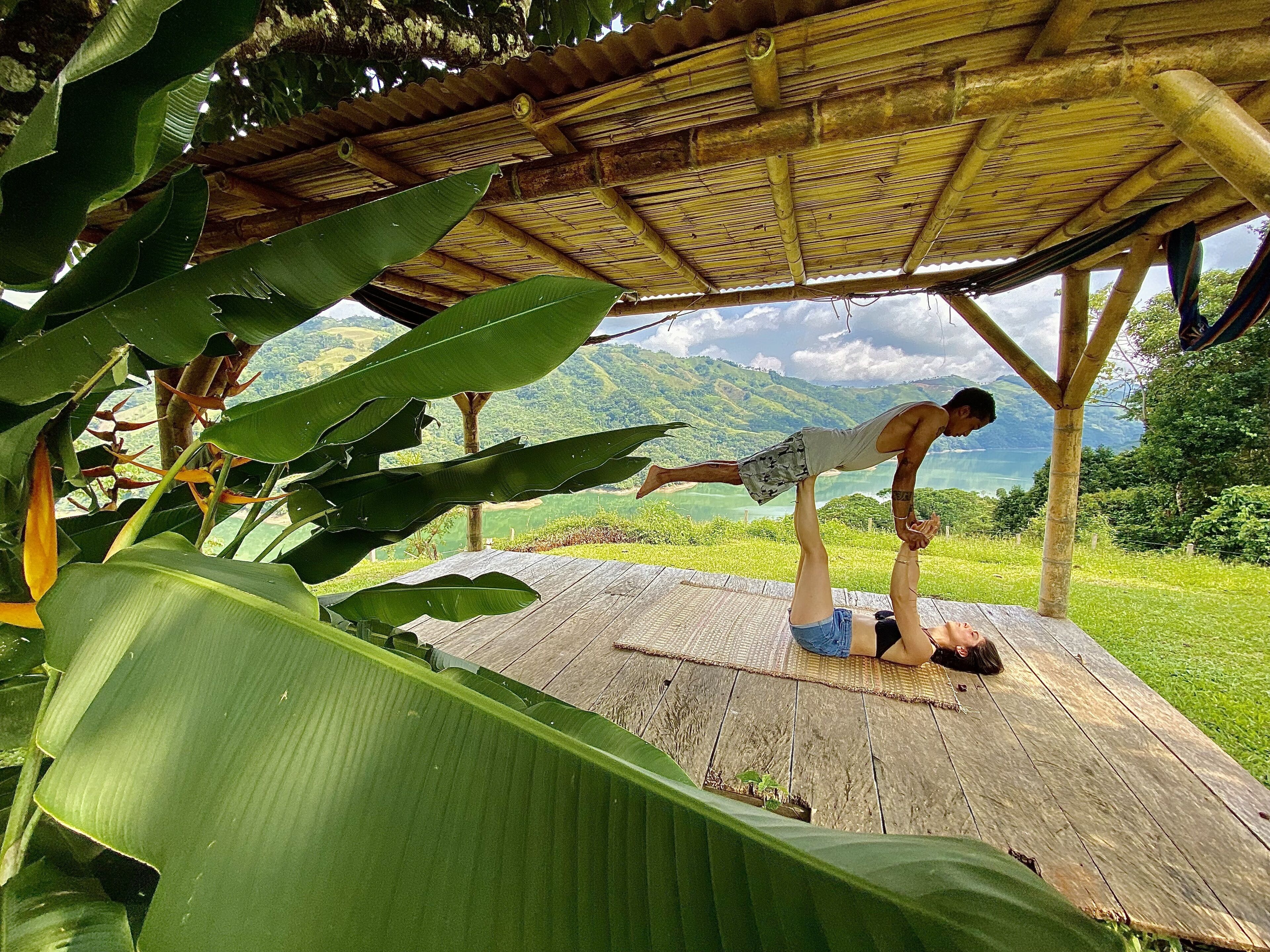A wooden deck with a bamboo roof, surrounded by lush greenery and a scenic view of mountains and a lake.