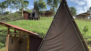 Un camping con una tienda de campaña triangular, un pequeño baño rústico con inodoro y algunas cabañas al fondo.