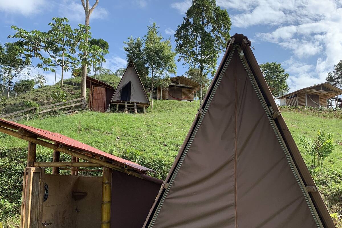 Un camping con una tienda de campaña triangular, un pequeño baño rústico con inodoro y algunas cabañas al fondo.