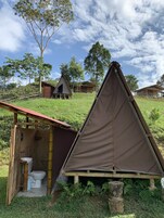 Un camping con una tienda de campaña triangular, un pequeño baño rústico con inodoro y algunas cabañas al fondo.