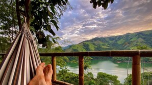 Una hamaca con los pies de una persona descansando sobre ella, con vistas a un valle verde y un lago.