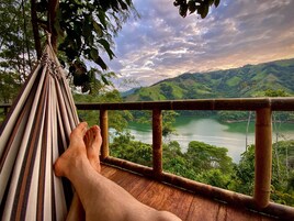 Una hamaca con los pies de una persona descansando sobre ella, con vistas a un valle verde y un lago.