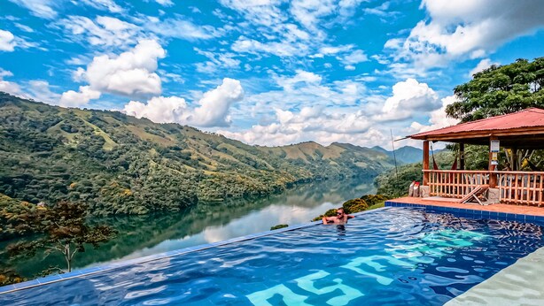 Una piscina infinita con vista a un lago y montañas, una terraza de madera y una estructura con techo rojo.